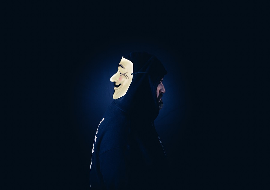 Silhouette d'une personne portant un sweat à capuche et un masque anonyme blanc stylisé, sur fond sombre pour présenter une usurpation d'identité.